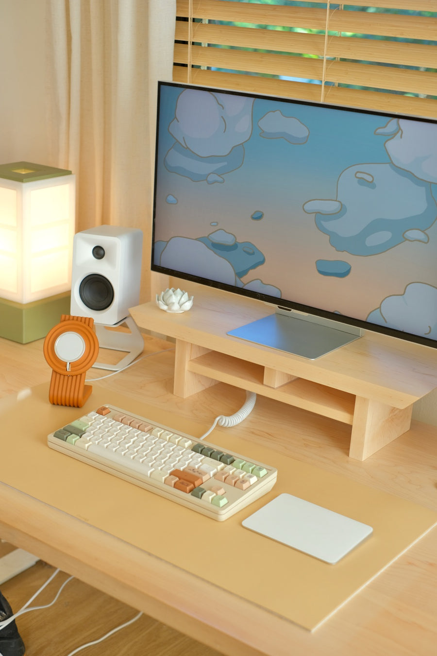 A monitor on a maple desk shelf showing the cloud wallpaper, above a keyboard and a Charger Stand on a desk mat