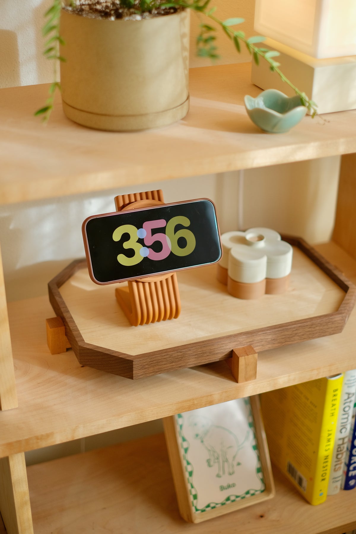 Coming this fall: the Zen MagSafe Charger Stand in Burnt Orange holding an iPhone sideways in standby mode, showing the clock, on a walnut Zen Tray beside a Fa Trinket Tray duo, on a bookshelf