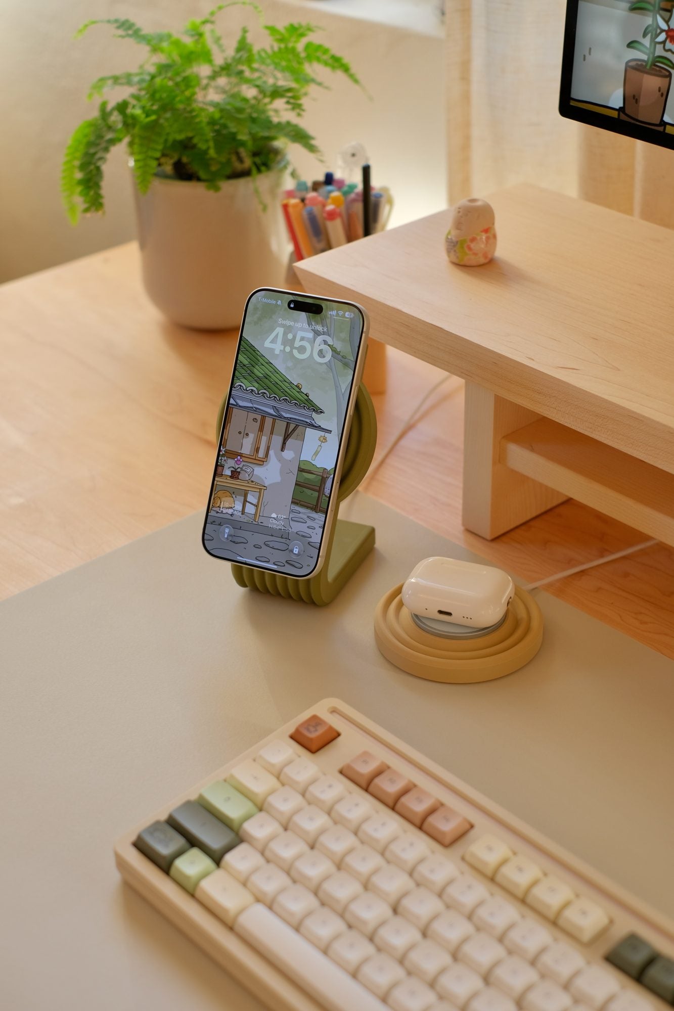 A Moss Zen MagSafe Charger Stand holding an iPhone, with AirPods on a Caramel Zen MagSafe Charger Puck, on a Beige Cozyleigh Desk Mat