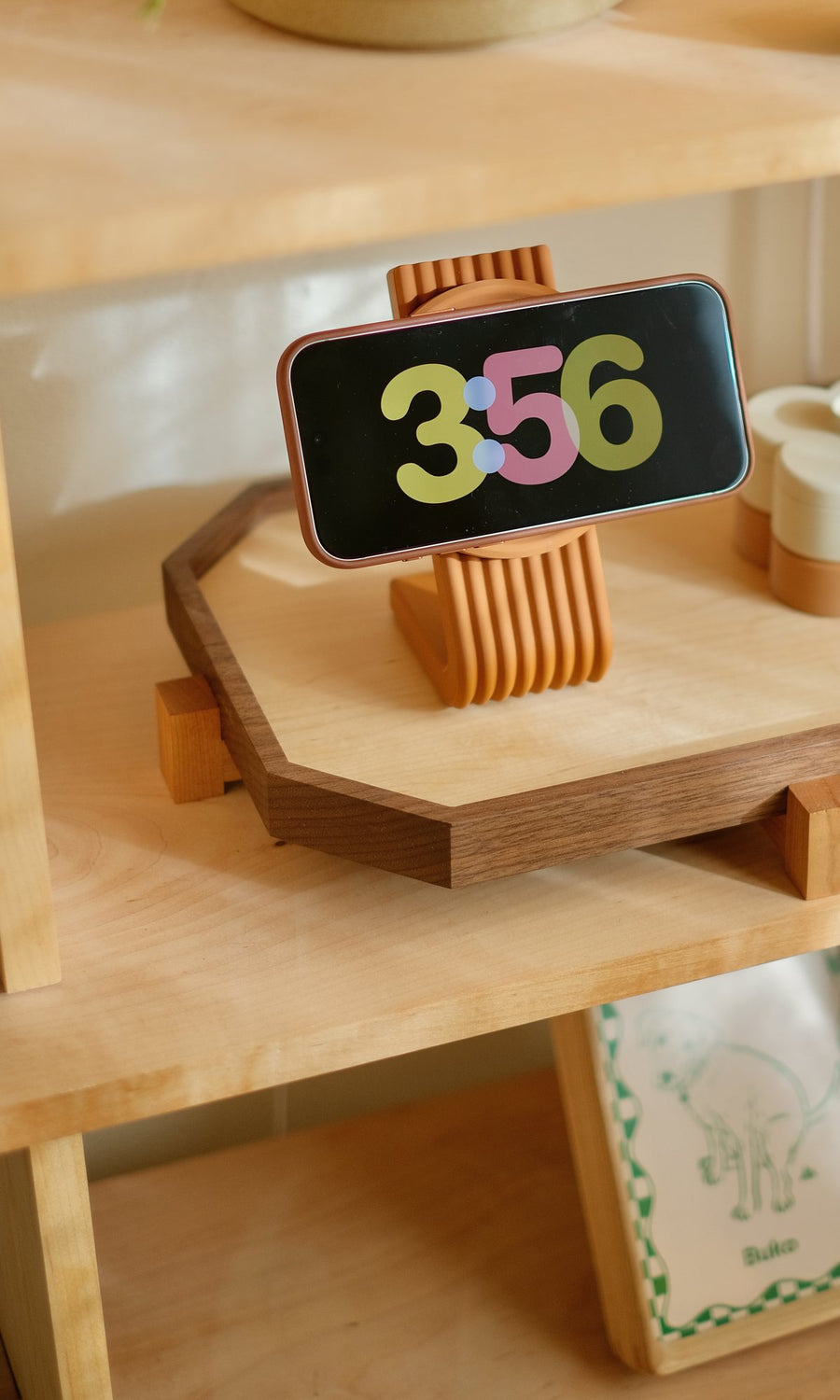 A phone on a Burnt Orange Zen MagSafe Charger Stand in standby mode, showing the clock, on a walnut Zen Tray on a maple bookshelf. Coming this fall in this colour.